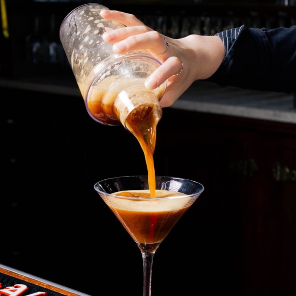 A bartender pours a frothy espresso martini from a shaker into a martini glass.