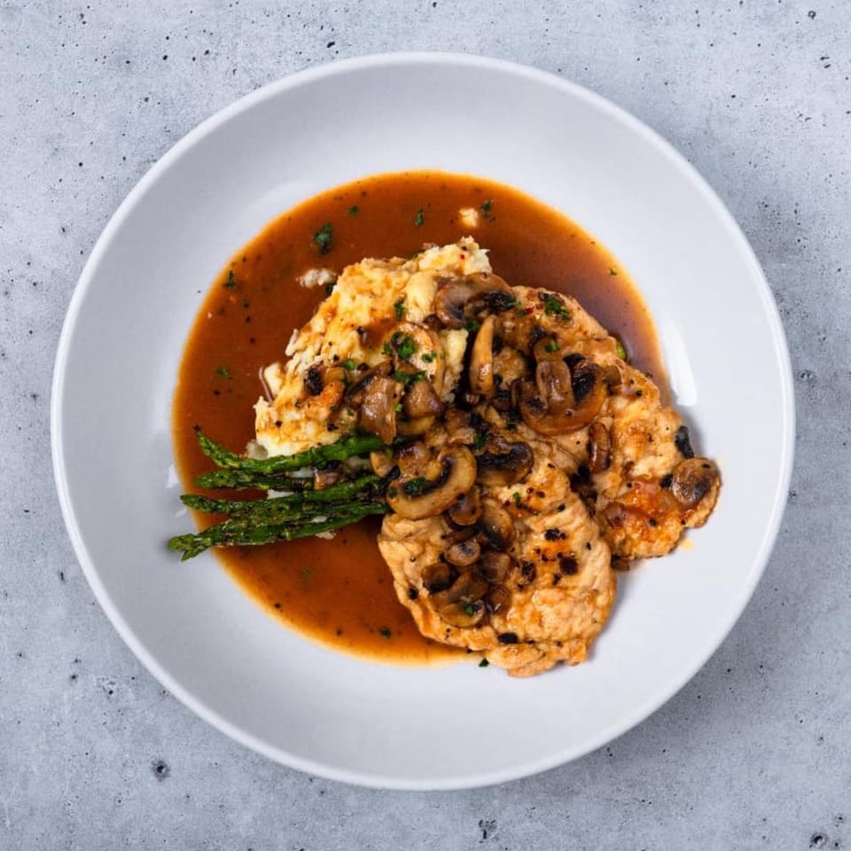 Chicken cutlets with mushrooms and asparagus in a rich brown sauce on a white plate.