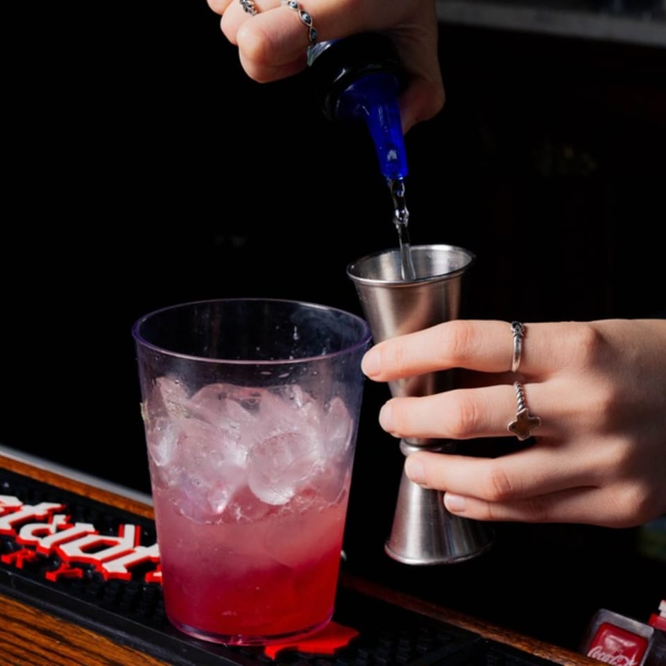 A bartender pours a clear liquid from a bottle into a jigger, next to a red iced drink.