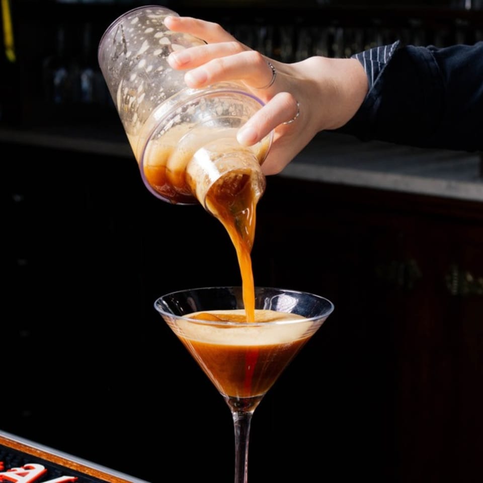 A bartender pours a frothy, dark amber espresso martini from a shaker into a martini glass.