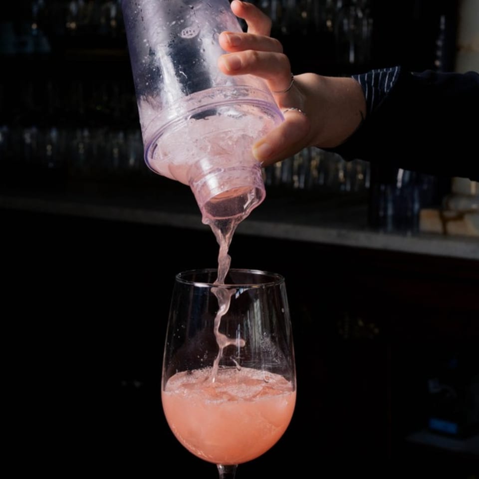 A bartender pours a pink cocktail from a shaker into a wine glass, creating a splash.
