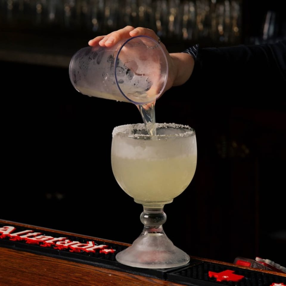 A bartender pours a margarita from a shaker into a large, salt-rimmed glass on a dark bar.