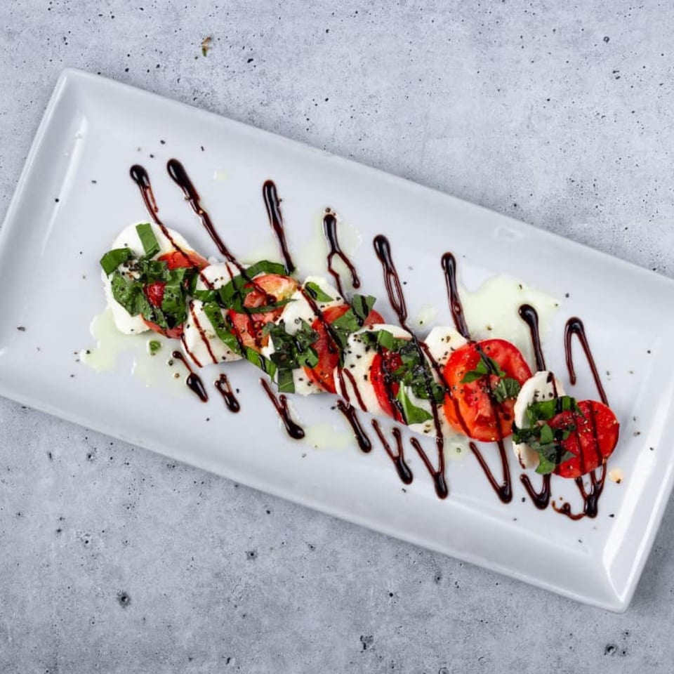 Caprese salad with sliced mozzarella, tomatoes, basil, olive oil, and balsamic glaze on a white plate.