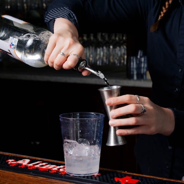 Bartender pouring clear liquid from a bottle into a jigger, with an ice-filled glass nearby.