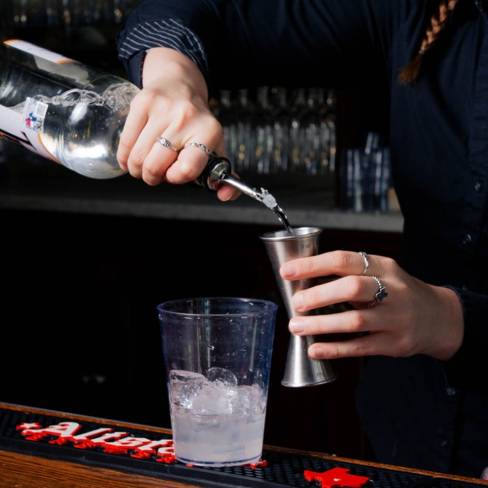 Bartender pouring clear liquid from a bottle into a jigger, with an ice-filled glass nearby.