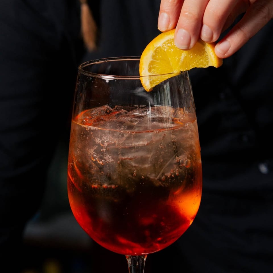 A hand garnishes a vibrant orange cocktail with ice and bubbles with a fresh orange slice.