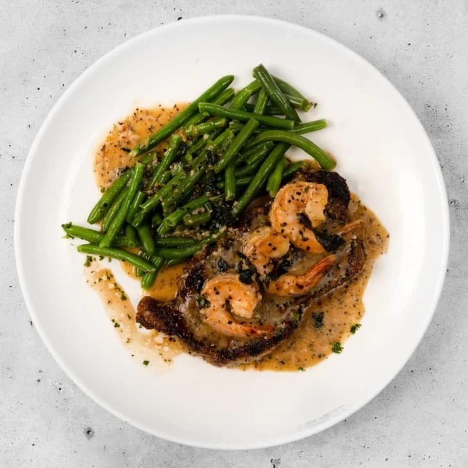 Steak and shrimp with green beans and a creamy sauce on a white plate.